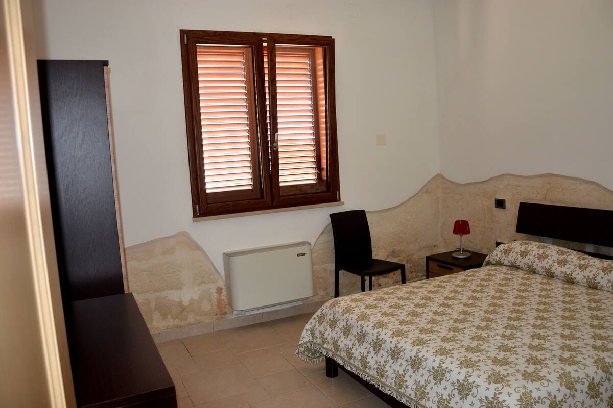 Une chambre à coucher comprenant un lit, une chaise, une table de chevet, un radiateur et une fenêtre avec des stores.