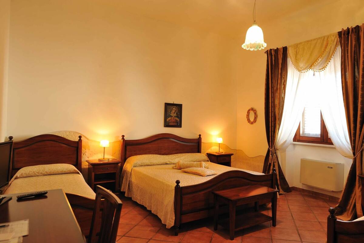 Une chambre d’hôtel avec deux lits simples, un bureau avec une chaise, une table de chevet avec une lampe, un miroir, un tableau encadré au mur et une fenêtre avec des rideaux.