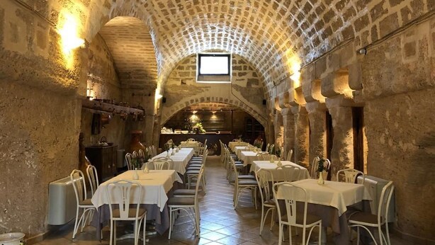 Ein Restaurant mit gewölbter Steindecke, langen Tischen und weißen Stühlen.
