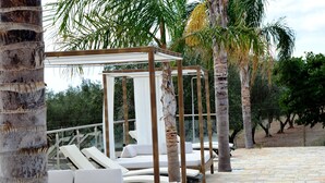 Ein überdachter Außenbereich mit Glastrennwänden, weißen gepolsterten Liegeloungern und einer Pergola.