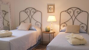 Deux lits simples avec des têtes de lit en métal, une petite table ronde avec une lampe et un tableau encadré au mur.