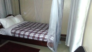 A single bed with a plaid bedspread and white pillows, a mosquito net, and a dark wooden headboard.