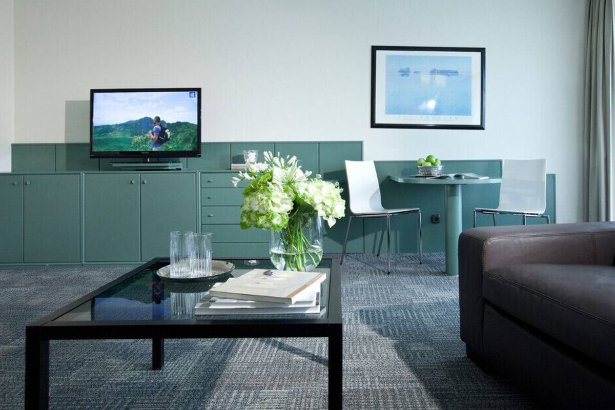 Ein modernes Hotelzimmer mit Flachbildfernseher, Schreibtisch mit Stuhl, Sofa, Couchtisch mit Blumenvase und einem gerahmten Bild an der Wand.