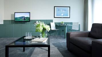 A modern hotel room with a flat-screen TV, a desk with a chair, a sofa, a coffee table with a vase of flowers, and a framed picture on the wall.