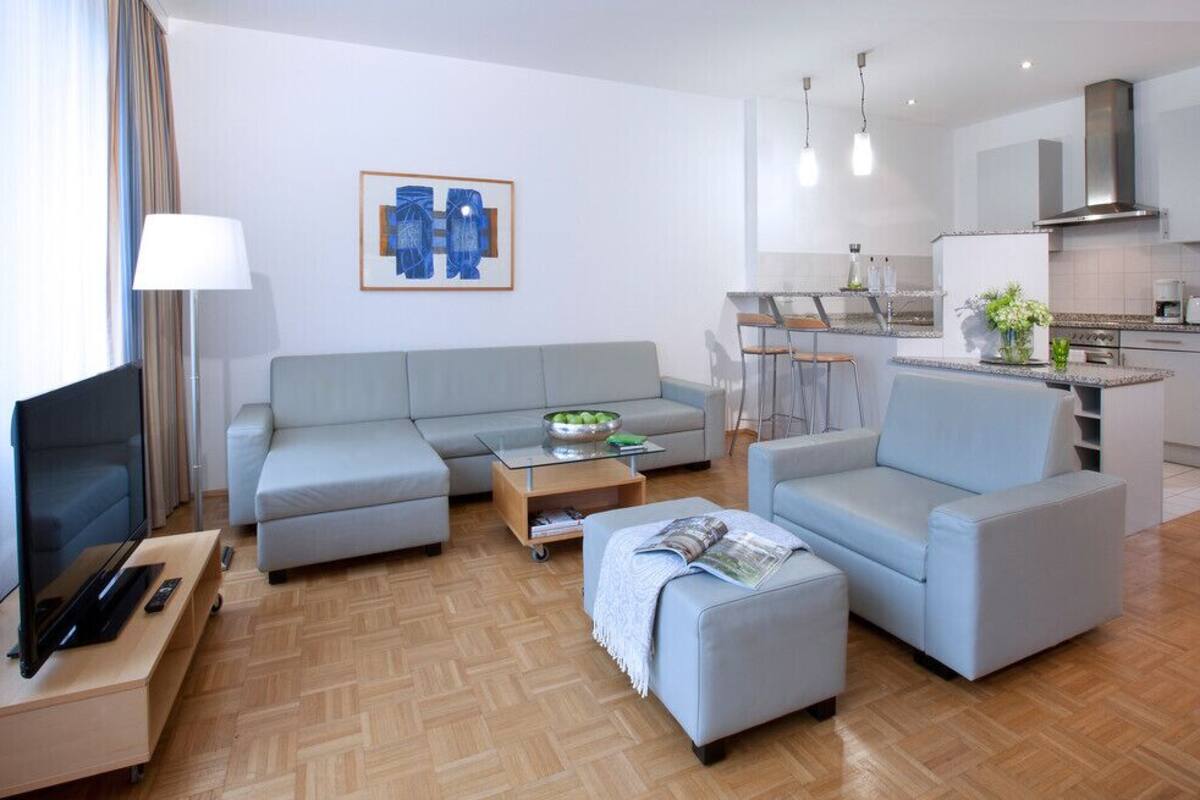 Ein modernes Wohnzimmer mit Sofa, Hocker und Couchtisch, einem Fernseher und einer Küchenzeile mit Esstisch.