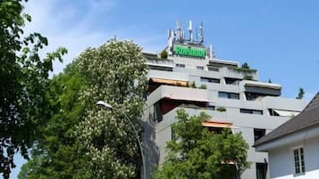 A modern hotel with a flat roof and multiple floors, surrounded by greenery and a clear sky.