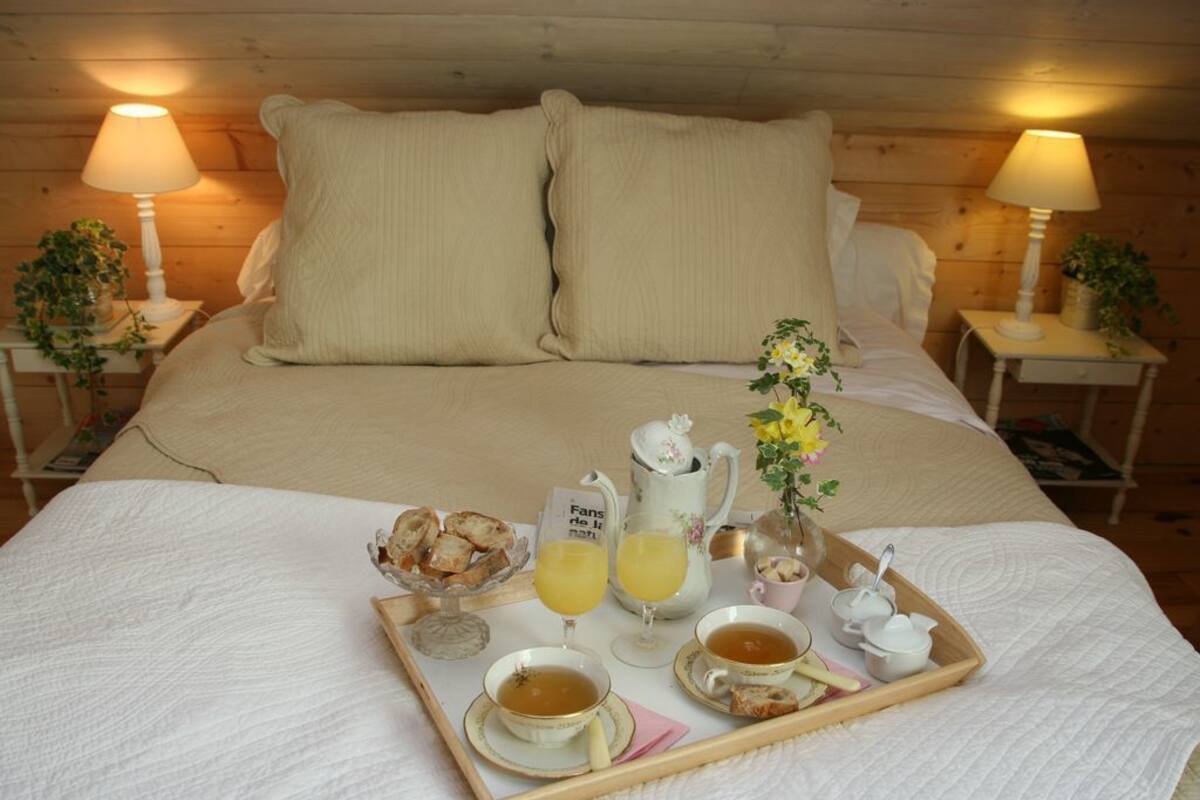 Un lit bien fait, avec un plateau contenant divers éléments pour le petit-déjeuner, notamment du thé, du jus de fruits et des viennoiseries, le tout placé devant un mur en bois et agrémenté de deux lampes.