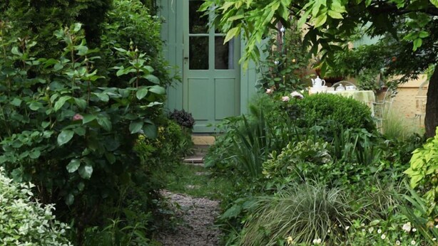 A garden pathway leading to a green door, surrounded by lush greenery and white flowers. - Chambres d’hôtes Au Jardin (Biscarrosse)