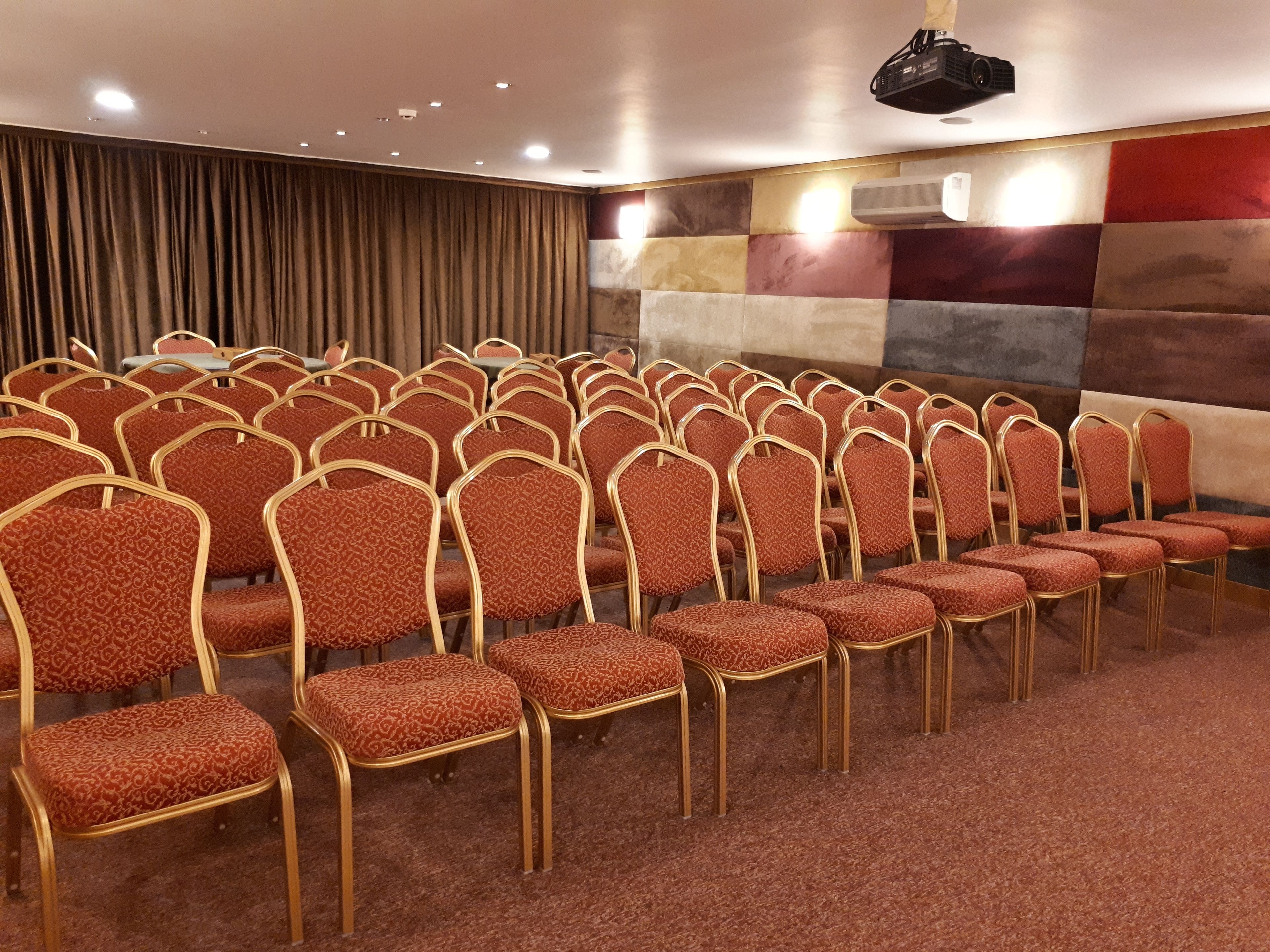 A conference room with rows of red upholstered chairs with gold frames, a carpeted floor, and a projector mounted on the ceiling.