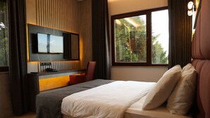 Habitación de hotel moderna con una cama grande, un televisor de pantalla plana montado en la pared, un escritorio con una silla y una ventana con vista a árboles.