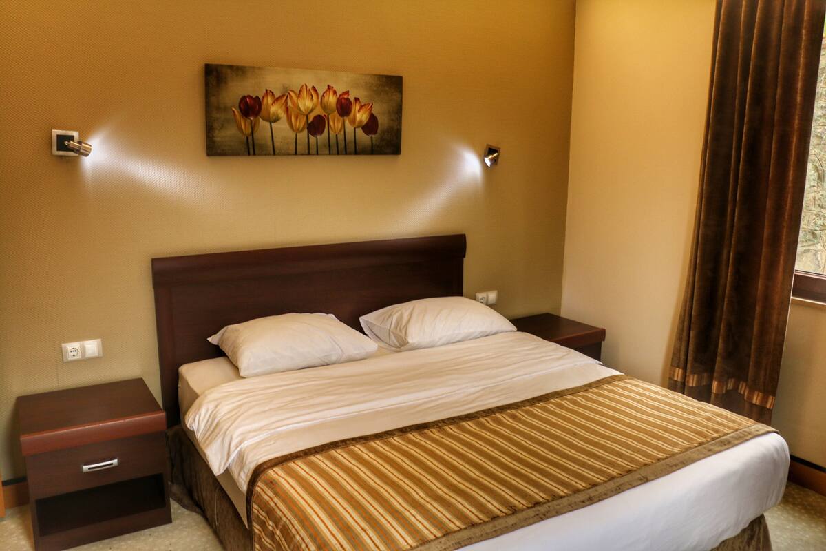 A hotel room with a bed, two nightstands, a painting of flowers, and a window with curtains.