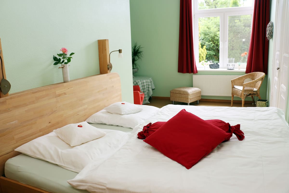 Una cama bien hecha con almohadas blancas y rojas, un cabecero de madera y una ventana con cortinas rojas.