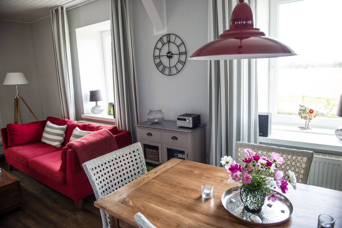 Una sala de estar acogedora con un sofá rojo, una mesa de comedor de madera con un jarrón de flores, un reloj en la pared y una ventana con cortinas.