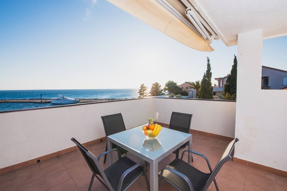 A balcony with a table and chairs, a fruit bowl, and a view of the sea and boats.