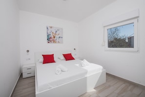 Een moderne slaapkamer met een groot bed, witte beddengoed, rode kussens, een klein nachtkastje, een wanddecoratie en een raam met uitzicht op bomen.