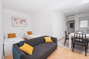 Een moderne, minimalistische woonruimte met een grijze bank, twee witte bedden en een eethoek.