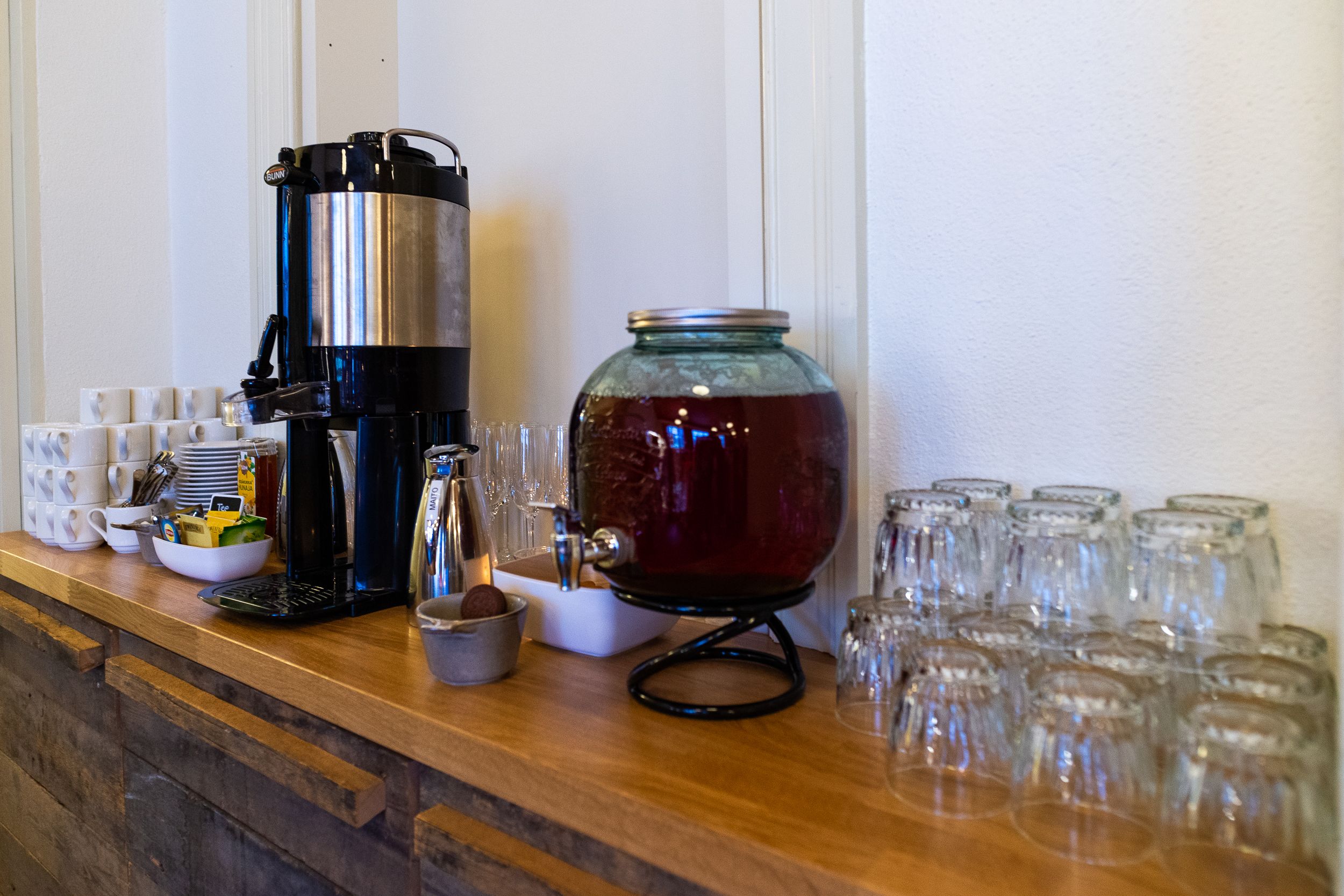 Uma estação de café com cafeteira, um grande dispensador de água e vários copos e taças.