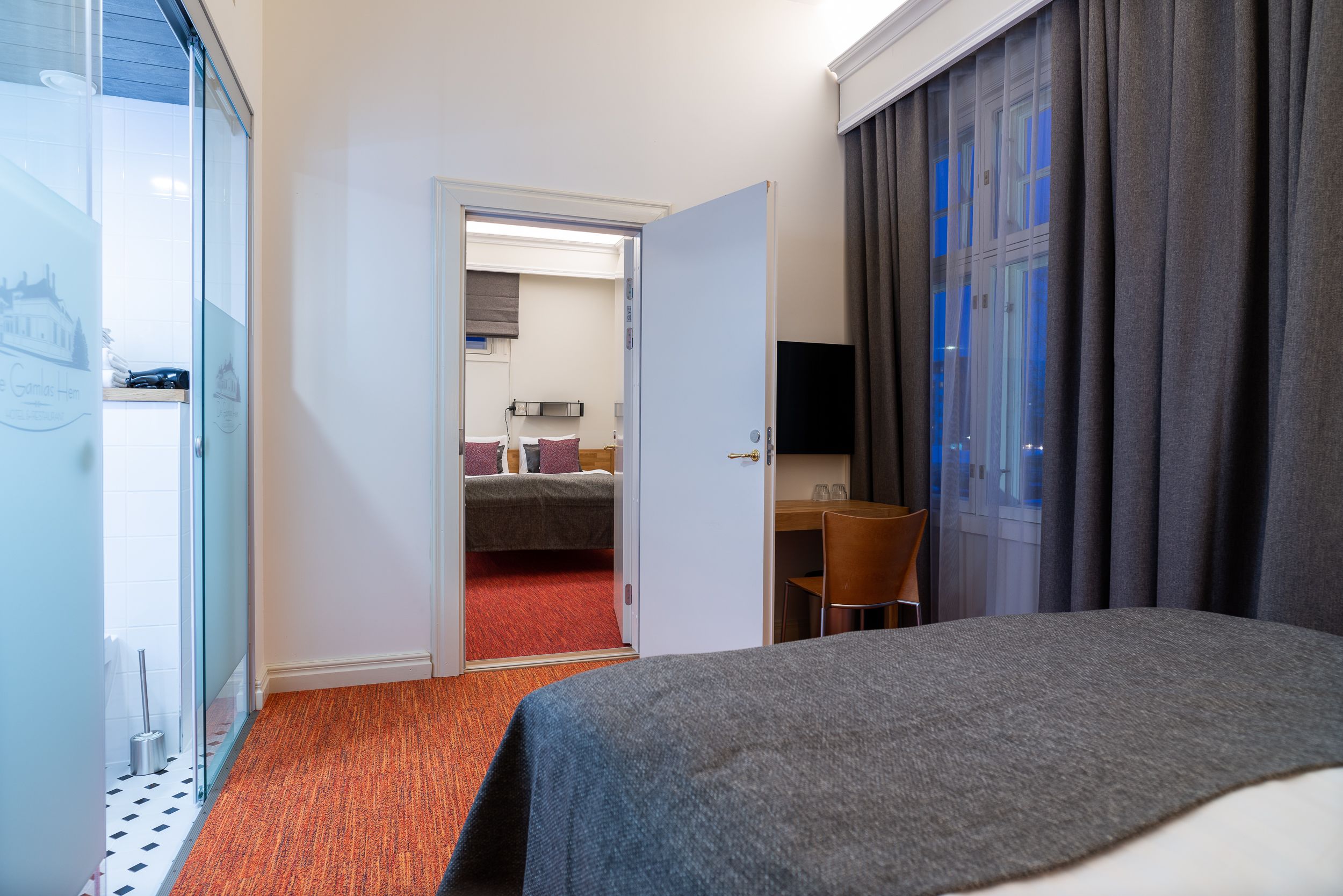 A hotel room with a bed, a desk, a chair, a TV, and a bathroom visible through an open door.