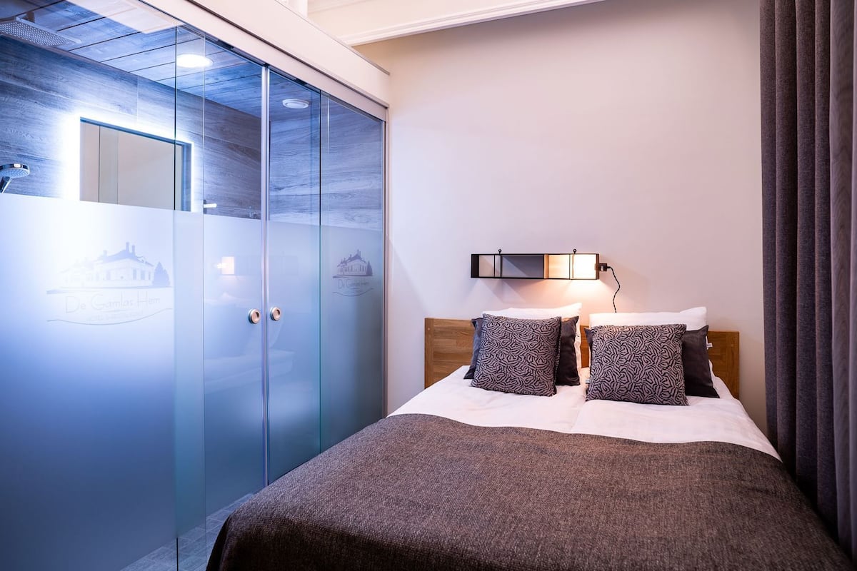 Un dormitorio con una cama, dos almohadones estampados, una mesita de noche, una lámpara de pared y un armario con puertas de vidrio esmerilado.