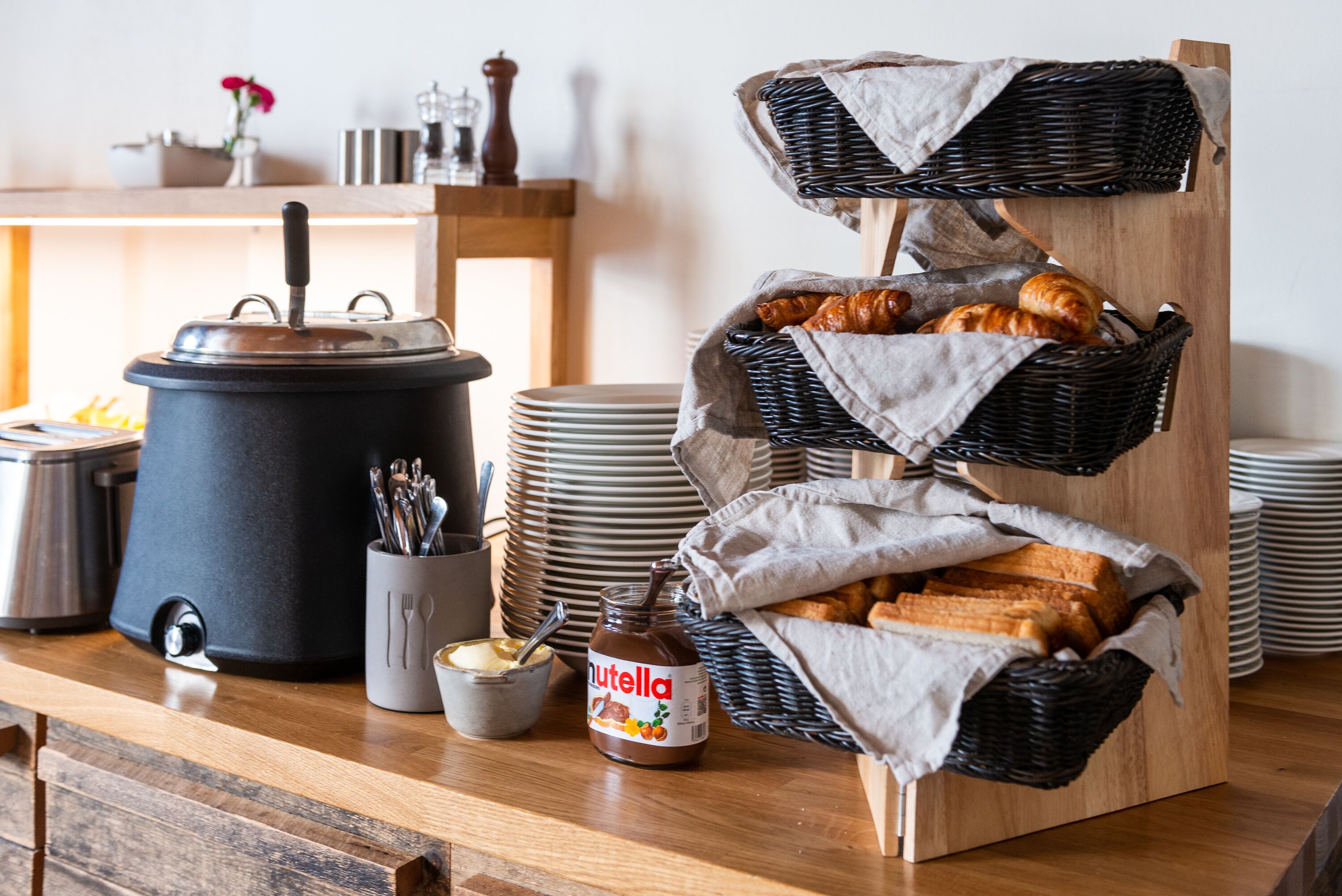 A kitchen counter with a slow cooker, a jar of Nutella, a basket of croissants, and stacked plates.