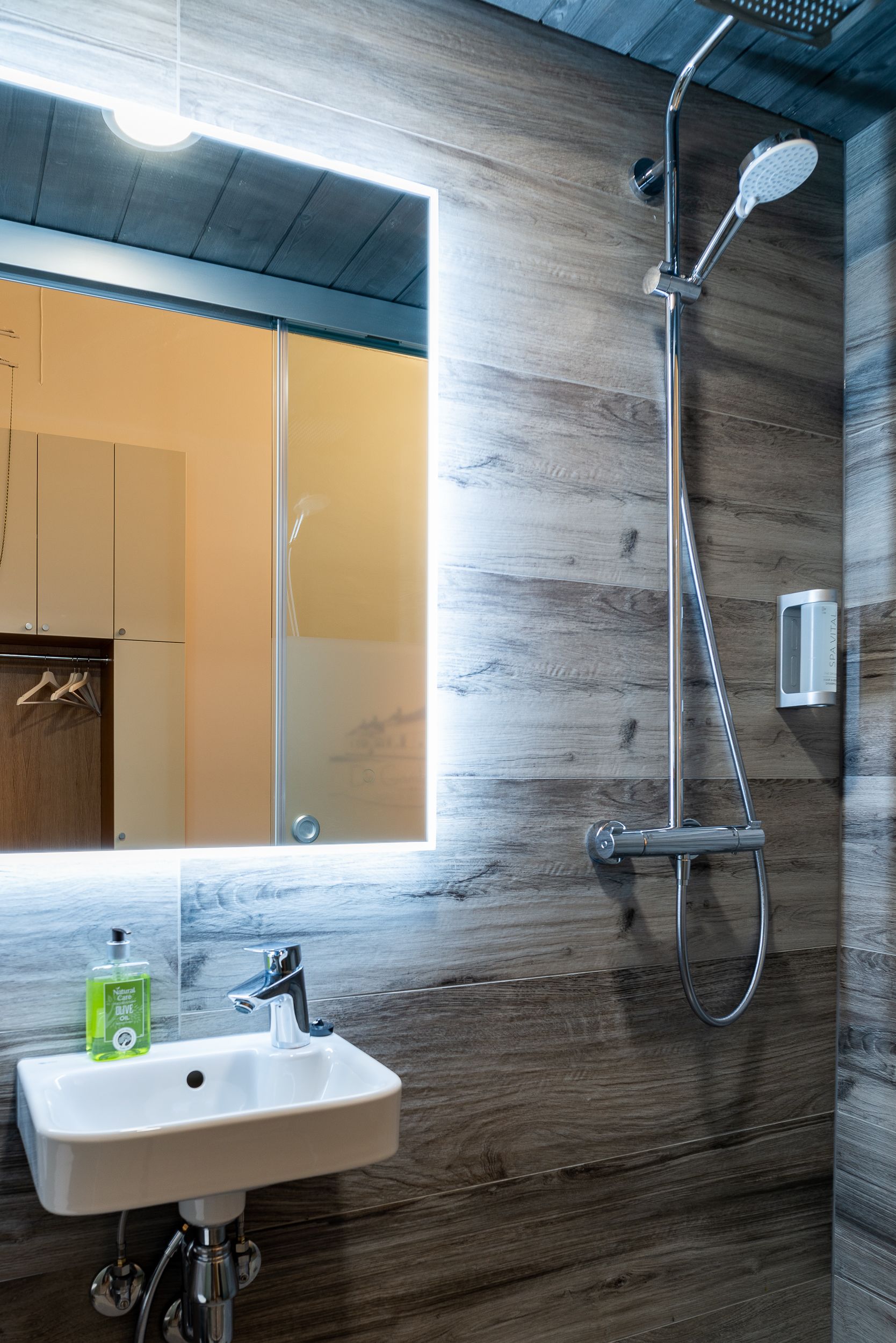 Un baño moderno con una pared de madera, un espejo grande, un lavamanos con dispensador de jabón y una ducha con cabezal de ducha manual.