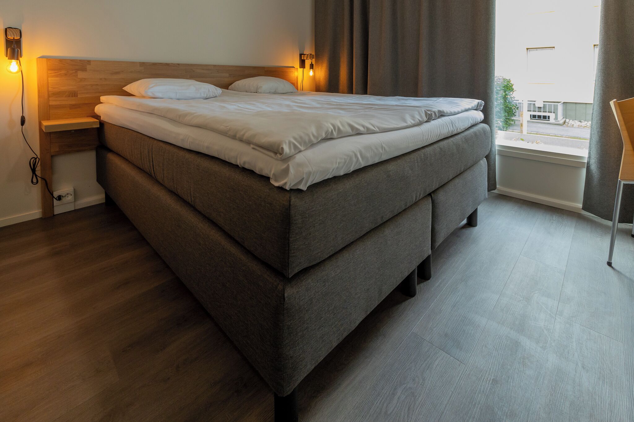A modern bedroom with a large bed, wooden headboard, bedside lamps, and a view of the outside through a window.