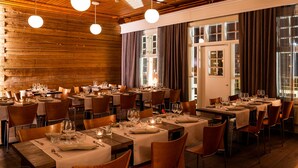 Un restaurante con varias mesas dispuestas para comer, paredes de madera y amplias ventanas con cortinas.