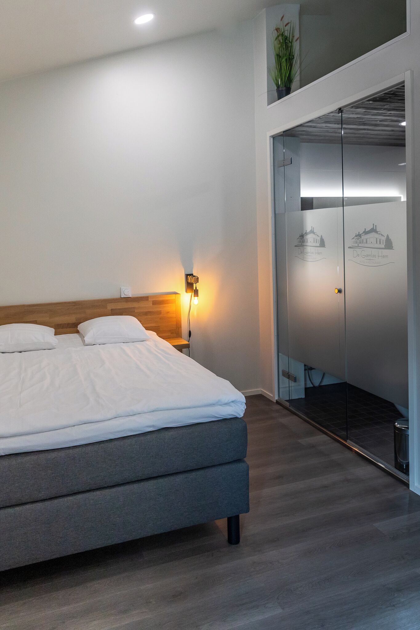 Une chambre d’hôtel moderne avec un grand lit, une table de chevet avec une lampe, une armoire à portes vitrées et une petite plante en pot accrochée au mur.