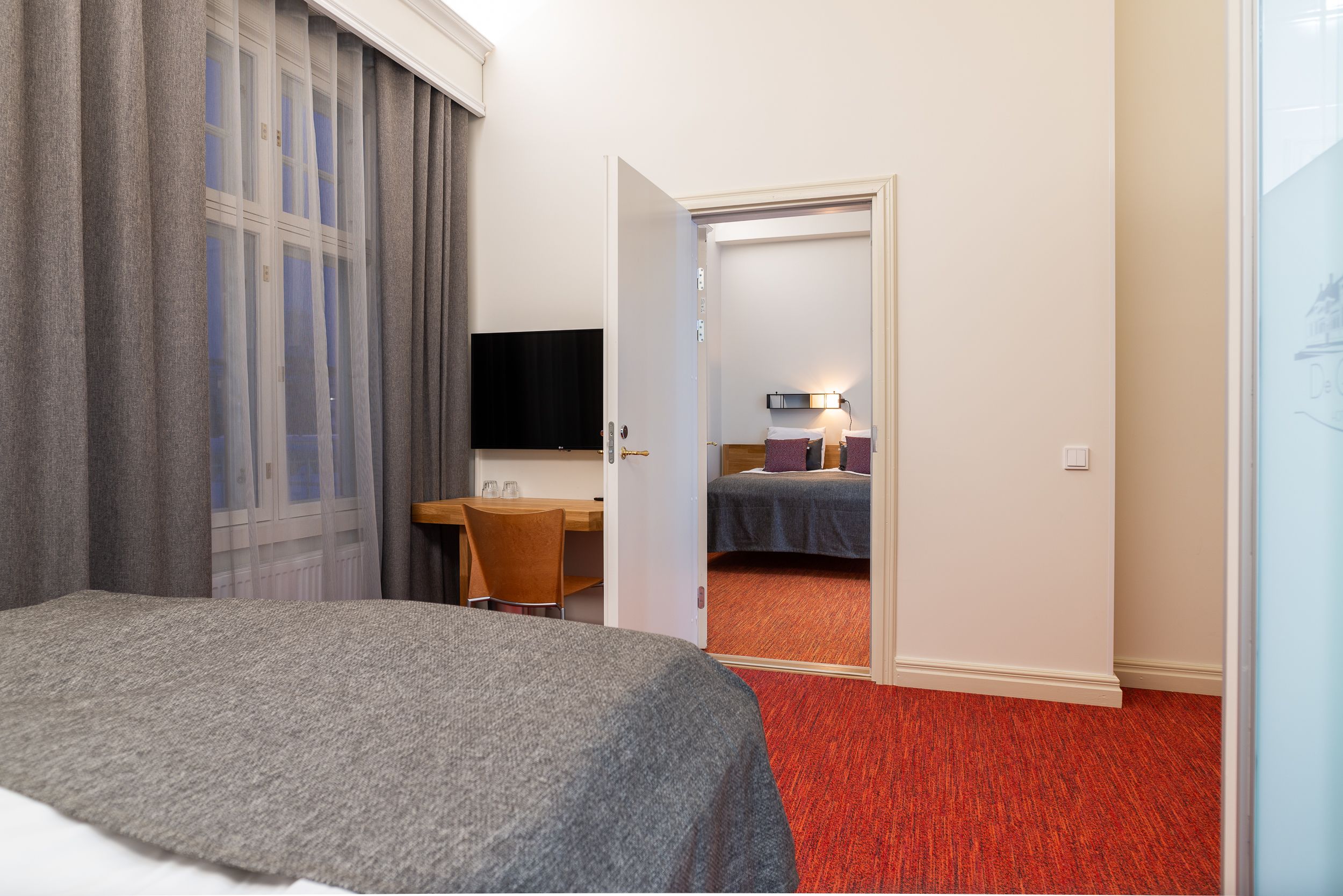 A hotel room with a bed, a desk, a chair, a television, and a door leading to another room.