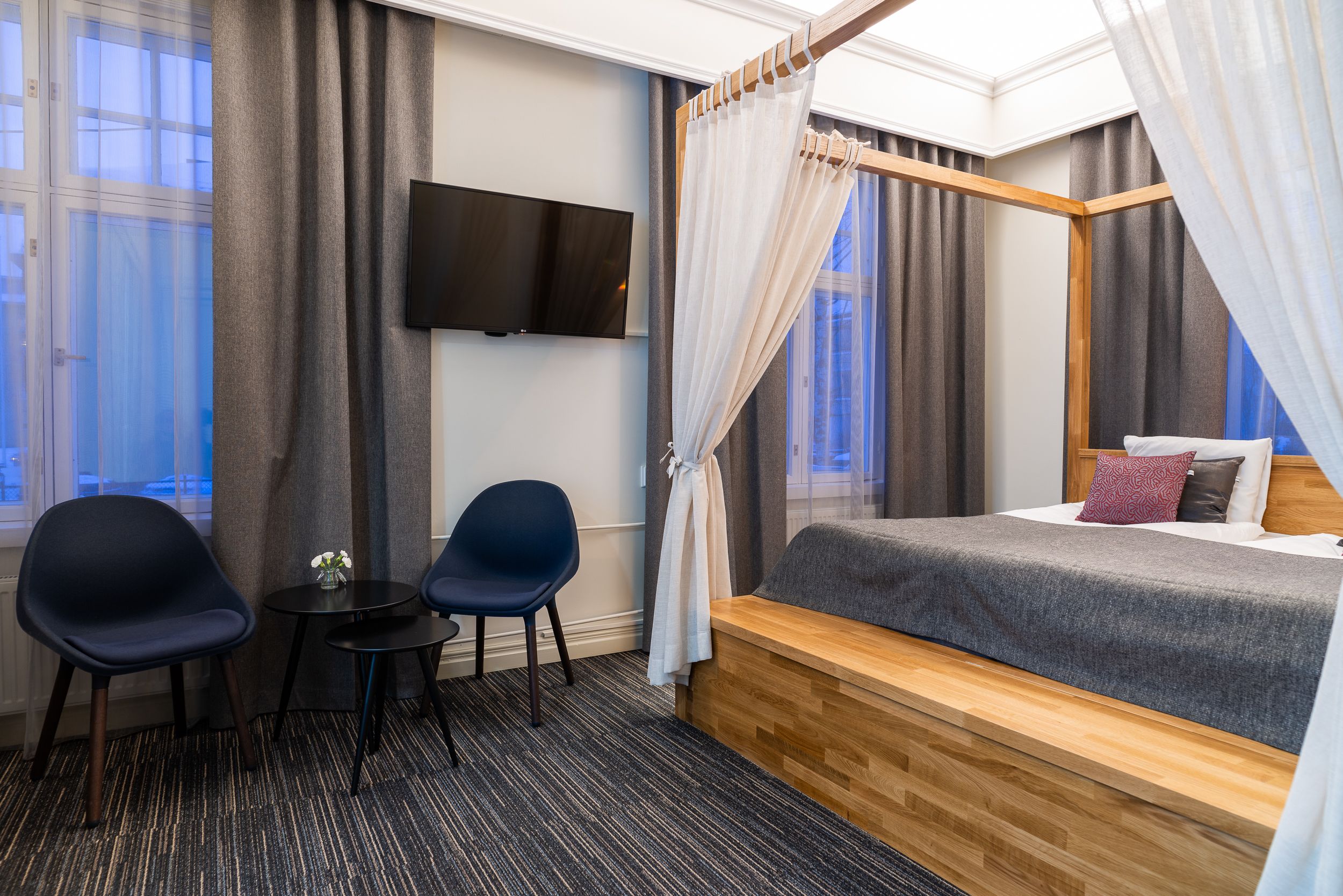 Ein Hotelzimmer mit Bett, Fernseher, zwei Stühlen, einem kleinen Tisch und einem Fenster mit Vorhängen.