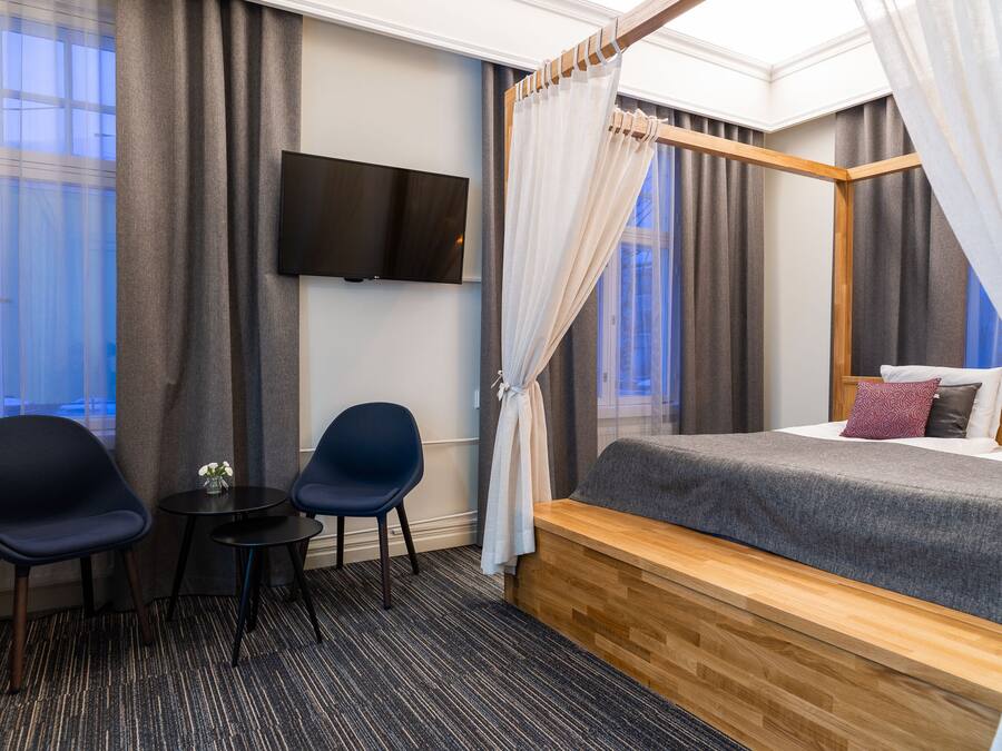 Ein Hotelzimmer mit Bett, Fernseher, zwei Stühlen, einem kleinen Tisch und einem Fenster mit Vorhängen.