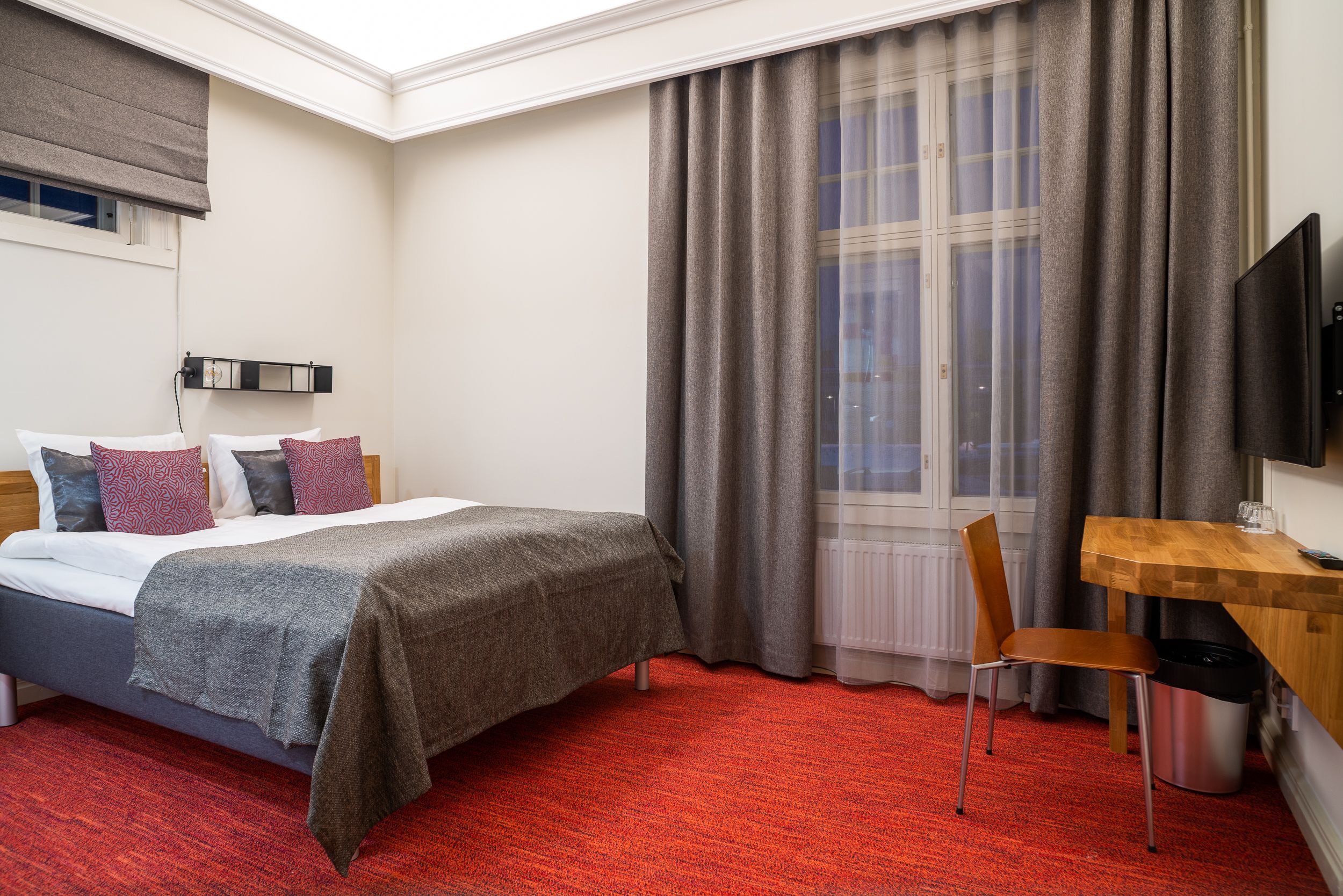 A hotel room with a bed, a desk, a chair, a TV, and a window with curtains.