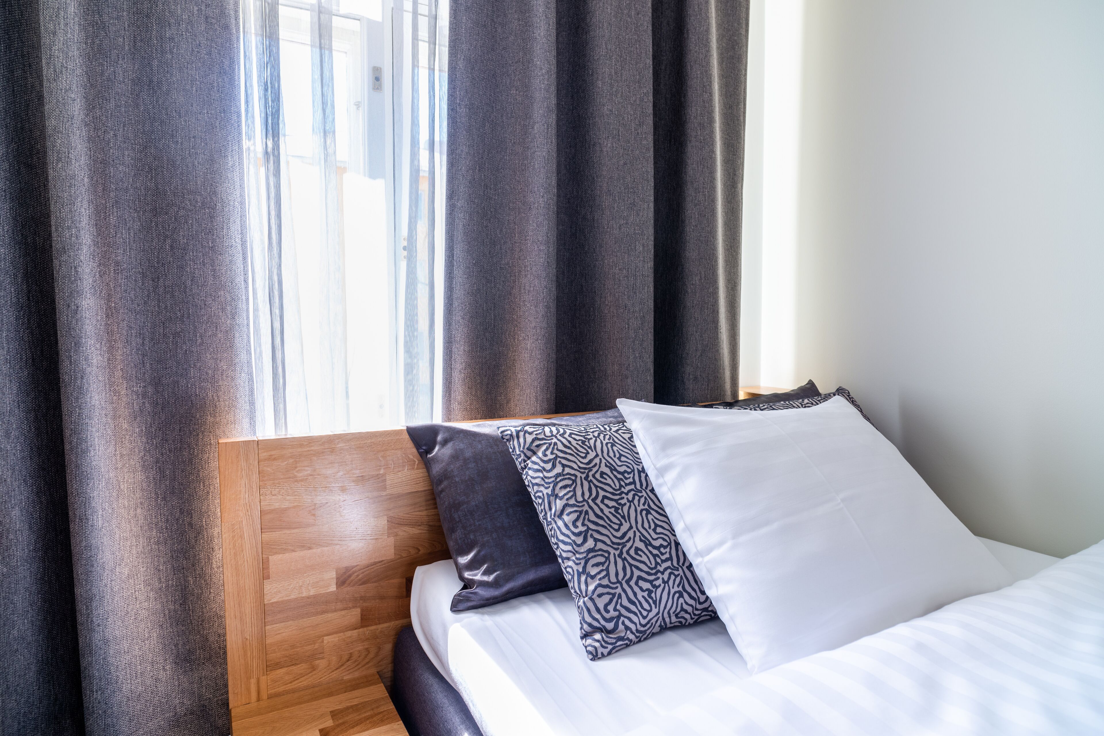 Una cama con cabecera de madera, colchón blanco y tres almohadas con distintos diseños.