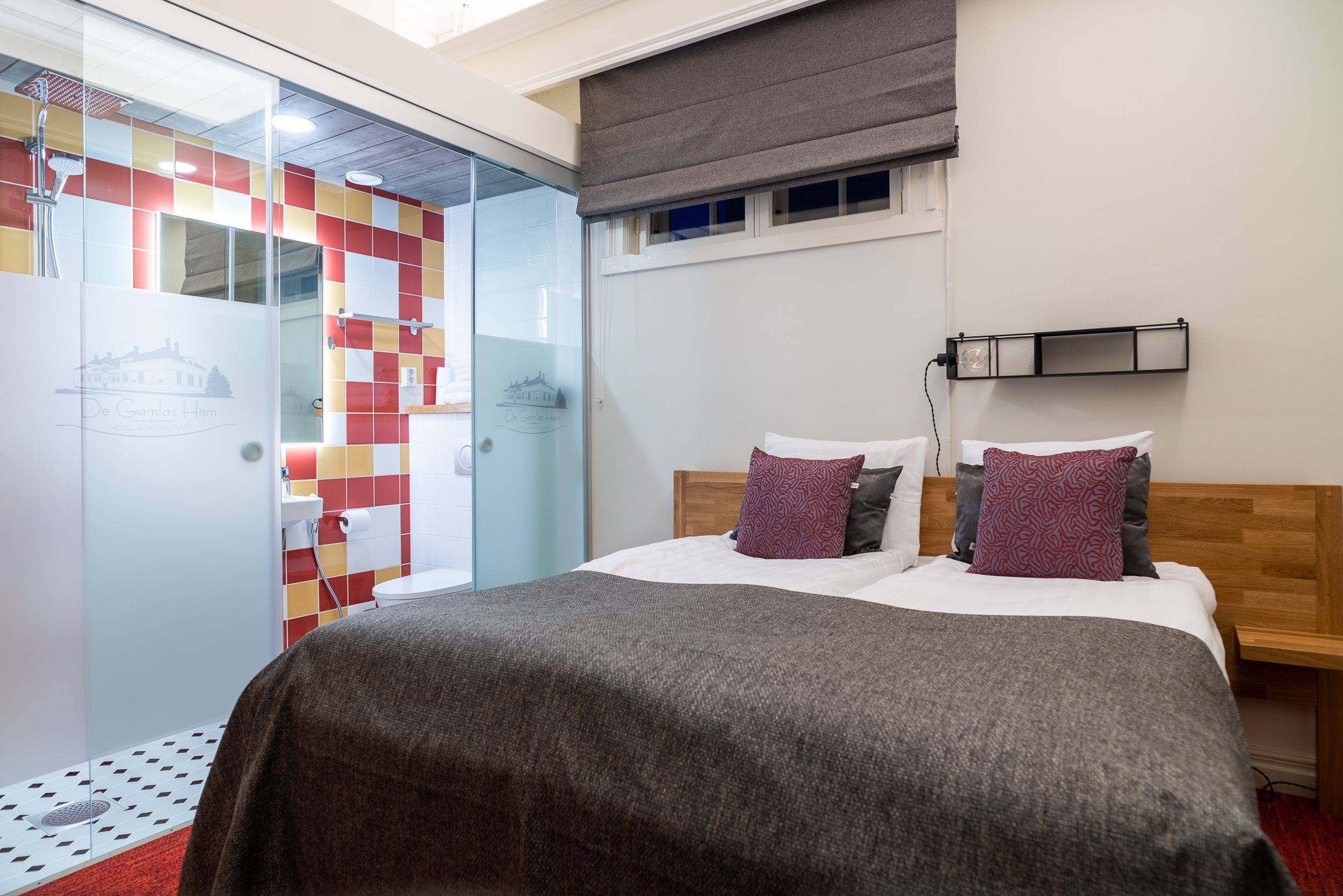 A hotel room with a bed, a bedside table, a nightstand, a window with a blind, and a bathroom with a shower and toilet.