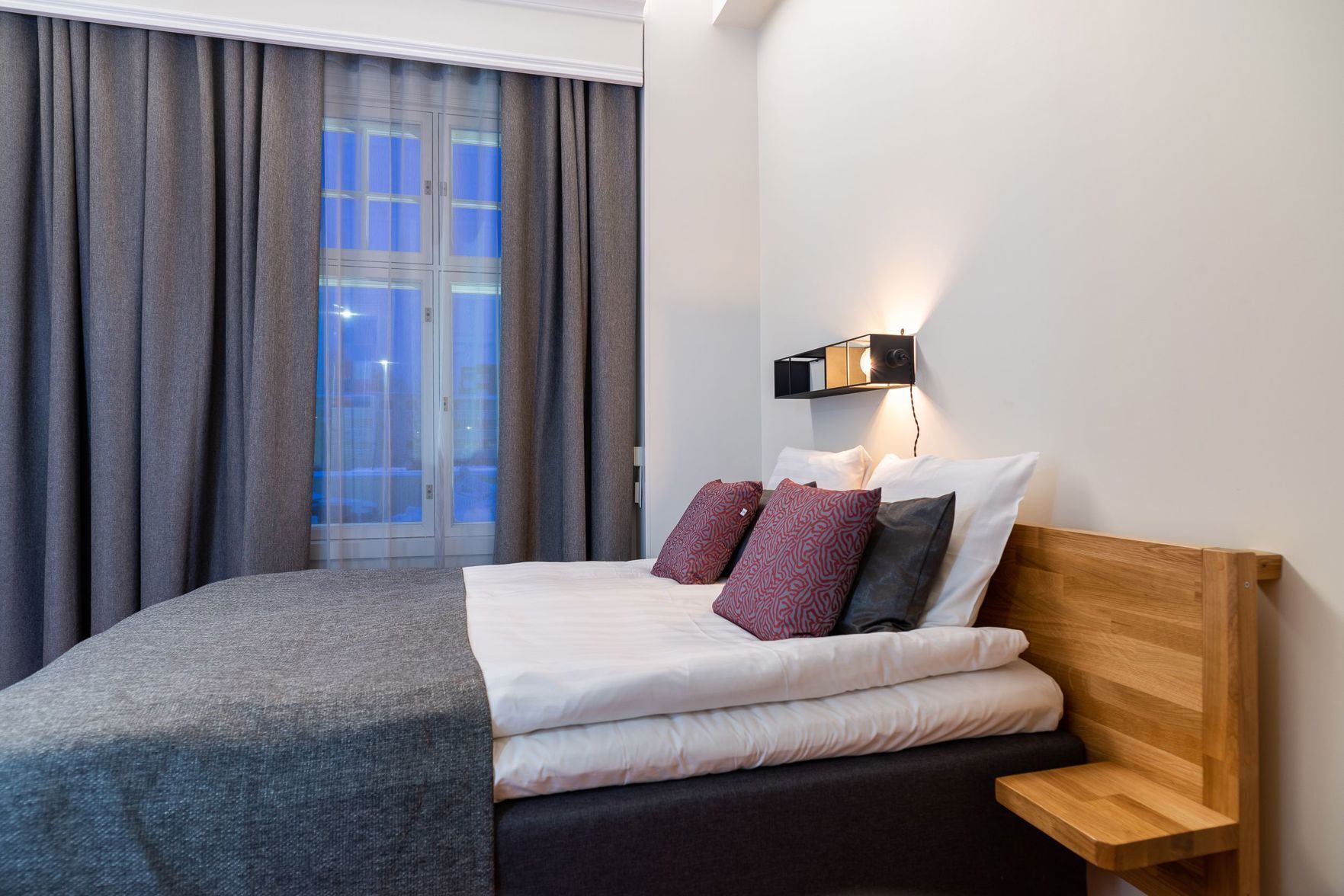 Ein Hotelzimmer mit Bett, Nachttisch, Wandleuchte und einem Fenster mit Vorhängen.