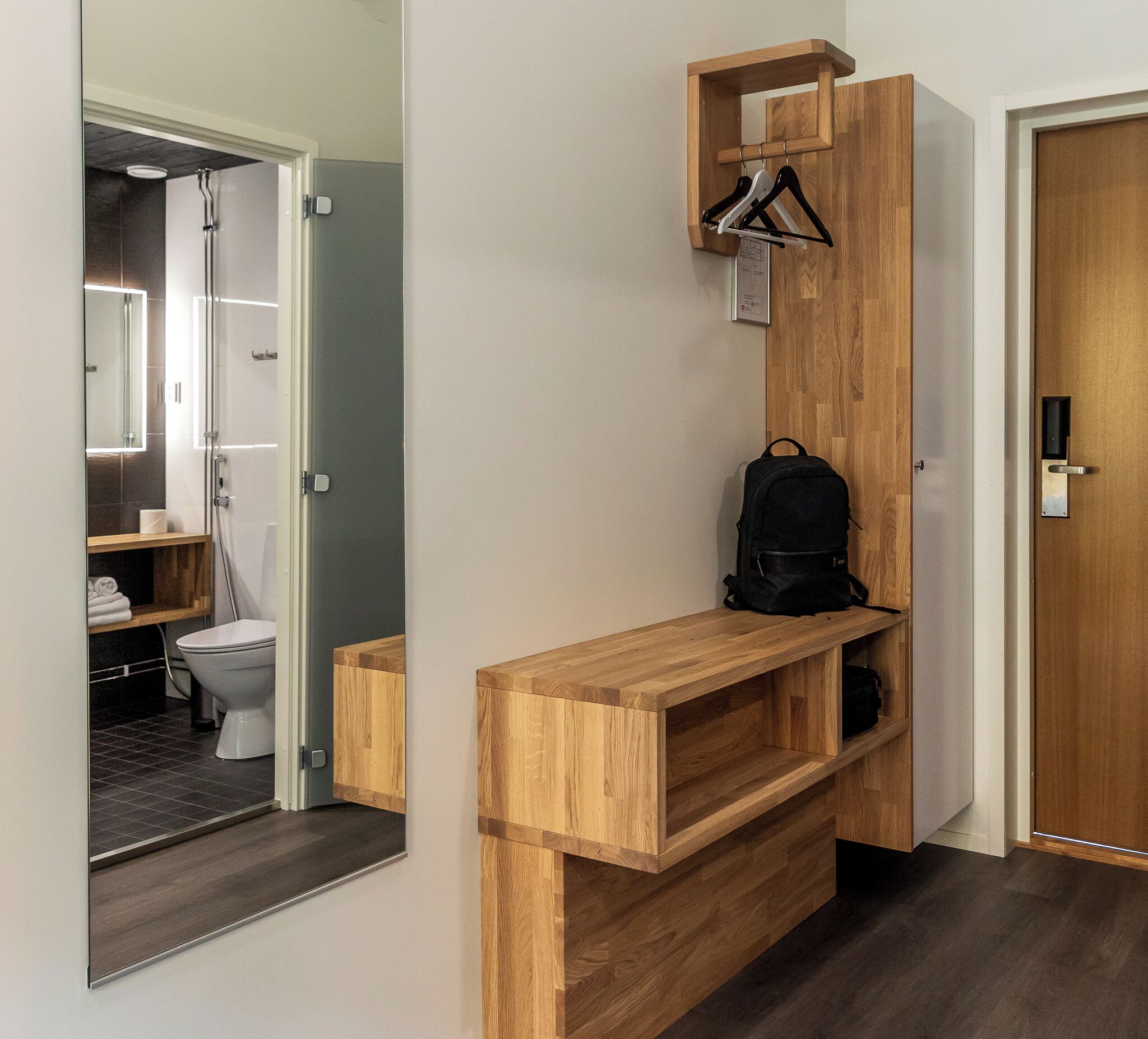 Un baño moderno con inodoro, lavamanos y ducha. Se observa un banco de madera con una mochila encima.