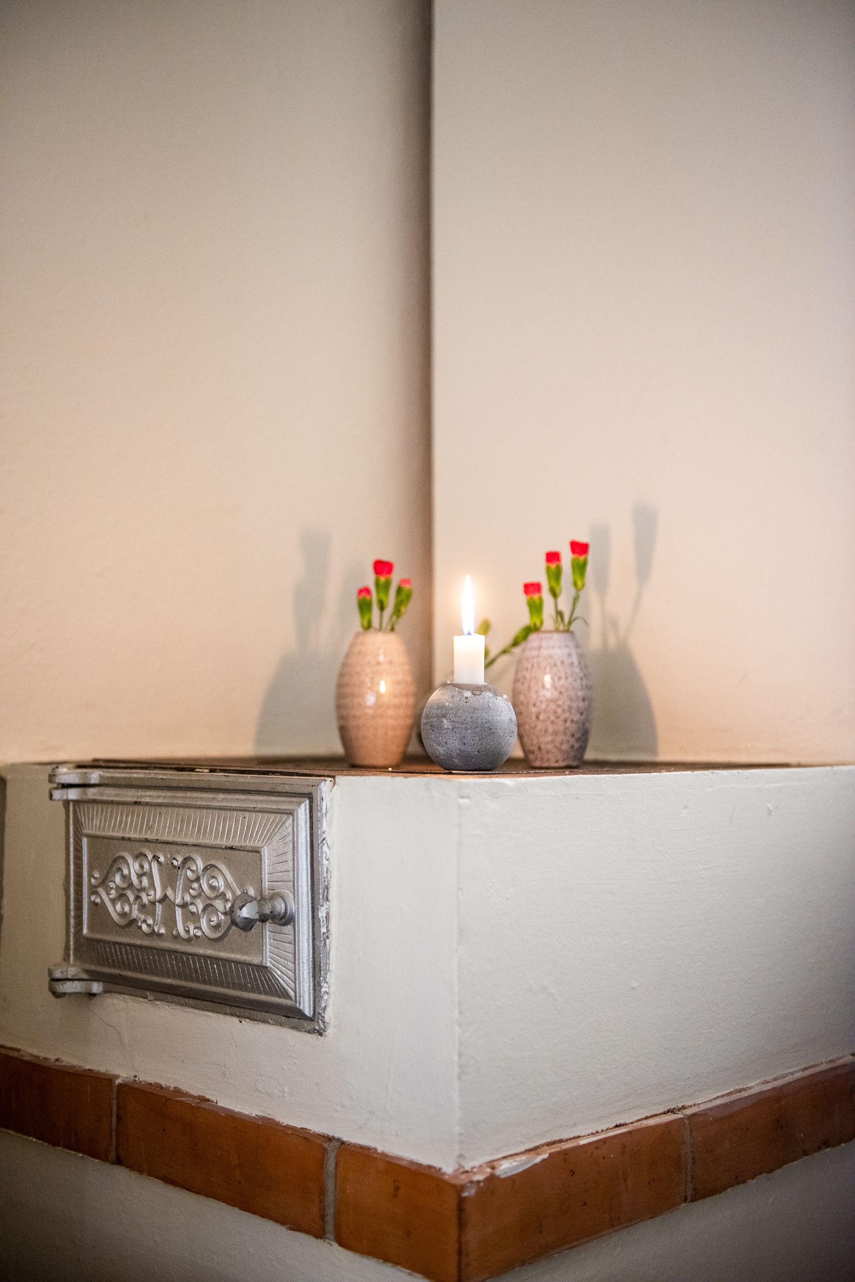 Un coin de pièce avec un mur blanc, une porte en métal dotée d’une poignée, et deux vases en céramique ornés de fleurs rouges et d’une bougie allumée.