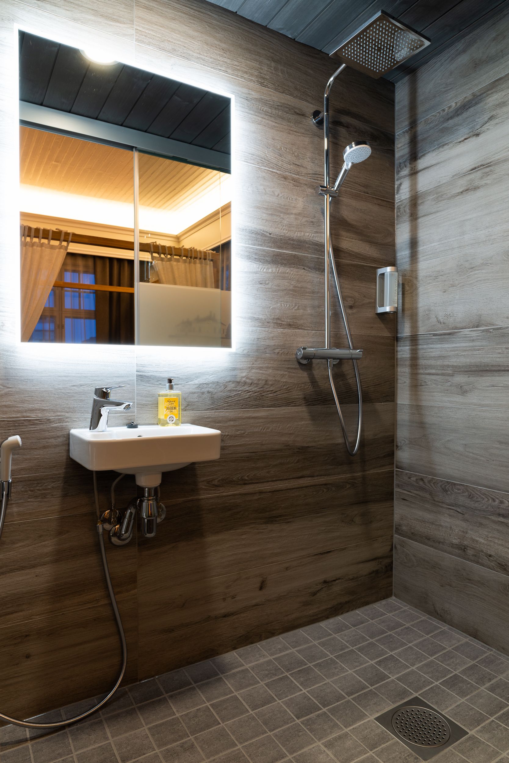 Un baño moderno con pared de madera, un espejo grande, un lavabo con dispensador de jabón y una ducha con cabezal de ducha de mano.