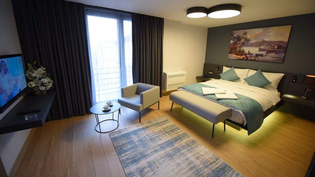 A modern hotel room with a bed, a desk with a TV, a chair, a small table, and a painting on the wall. - Stil Suit (Istanbul)