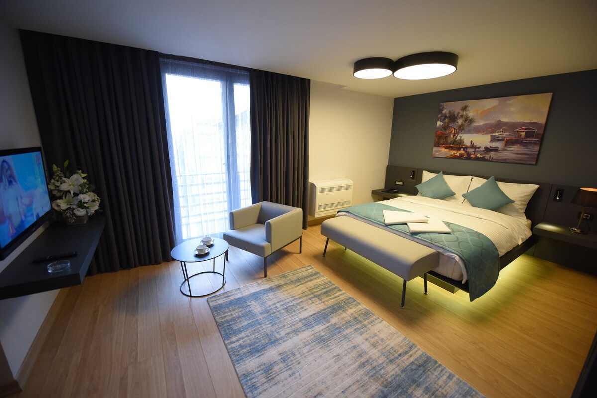 A modern hotel room with a bed, a desk with a TV, a chair, a small table, and a painting on the wall.