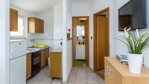 Cocina compacta con armarios de madera, un refrigerador blanco, una estufa y un lavamanos. Un pequeño pasillo conduce a un baño con un espejo y una planta en maceta.