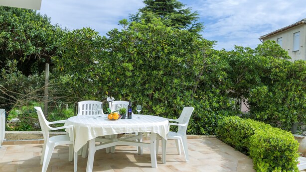 Un patio con una mesa y sillas blancas, rodeado de follaje y un piso de baldosas.