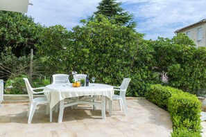 Une terrasse avec une table et des chaises blanches, entourée de verdure et dotée d’un sol carrelé.