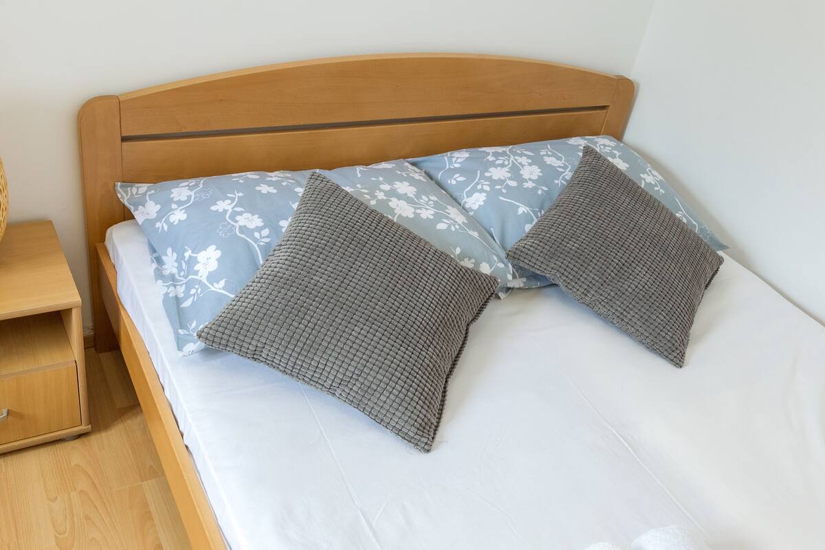 Una cama con cabecera de madera, dos almohadas con motivos florales y una almohada gris con textura.