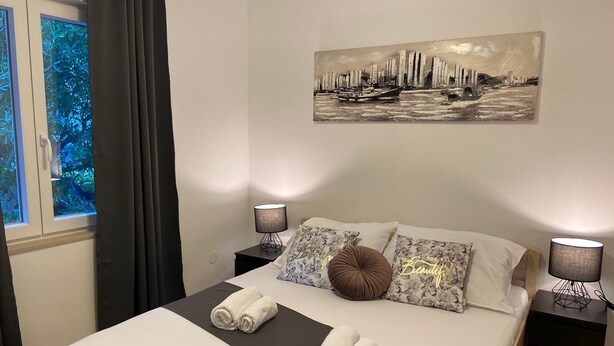 Cama bem arrumada com lençóis brancos e castanhos, duas luminárias de mesa, um quadro de paisagem urbana e uma janela com cortinas.