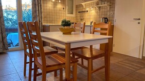 Uma sala de jantar com uma mesa branca e cadeiras de madeira, um vaso com planta, e uma cozinha ao fundo com paredes em azulejo e prateleiras.