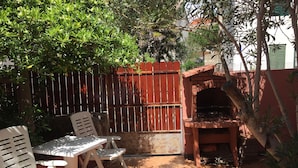 An outdoor patio with a brick floor, a wooden grill, and white plastic chairs. - Holiday Home Spektar (Novalja)