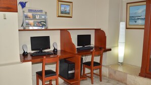 A computer lab with two desks, each with a computer, keyboard, and mouse. There is a monitor on the wall, a framed picture, and a sign for 'Correos del Ecuador'. - Grand Hotel Americano (Machala)