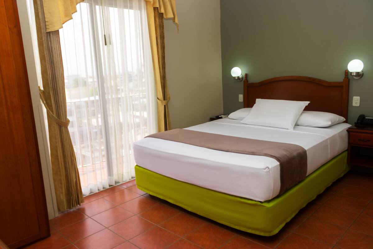 Una habitación de hotel con cama, cortinas, mesita de noche y lámpara de pared.