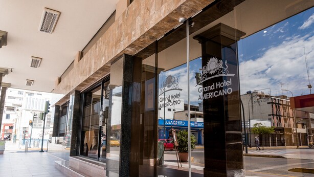 A hotel entrance with a glass facade, a prominent logo, and a street view outside. - Grand Hotel Americano (Machala)